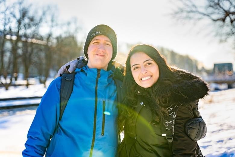Kaksi nuorta hymyilee talvisessa kaupunkimaisemassa. Aurinko paistaa.