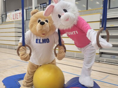 Elmo-nalle ja Viuhti-jänis Lietohallin liikuntasalissa tassut liikuntarenkaissa, lattialla värikkäitä jumppapalloja.