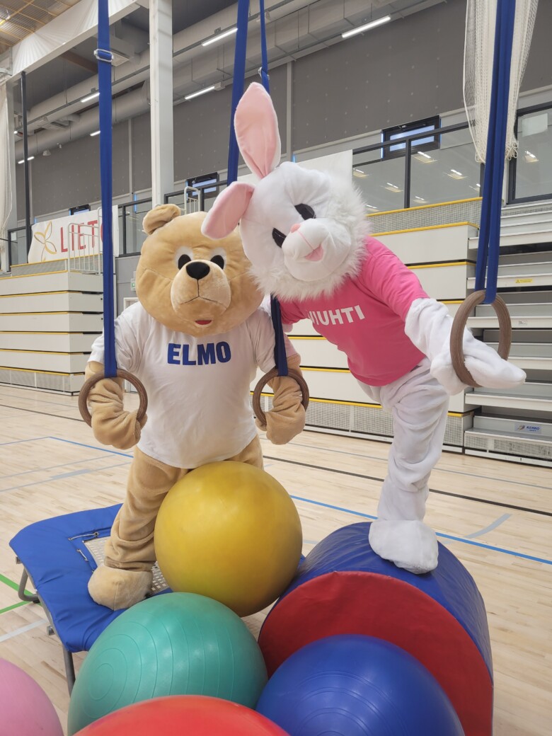 Elmo-nalle ja Viuhti-jänis Lietohallin liikuntasalissa tassut liikuntarenkaissa, lattialla värikkäitä jumppapalloja.