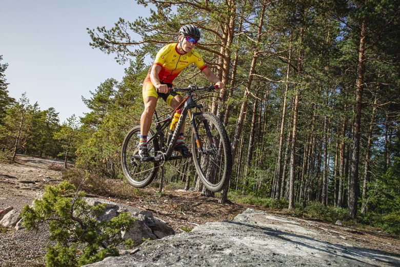 Maastopyöräilija ilmalennossa kallioisessa metsämaisemassa.