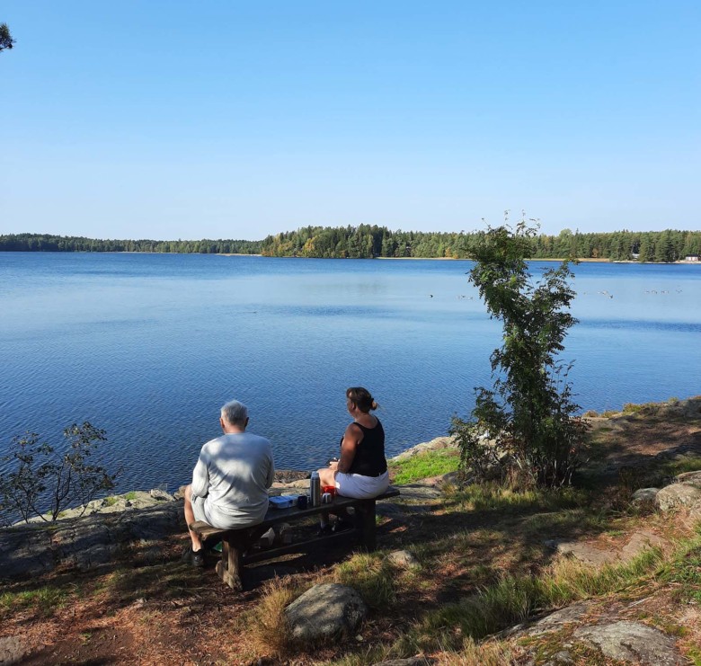 Kaksihenkilö istuu penkillä Littoistenjärven rannalla ja nauttii eväitä on kesä ja aurinkoinen sää.