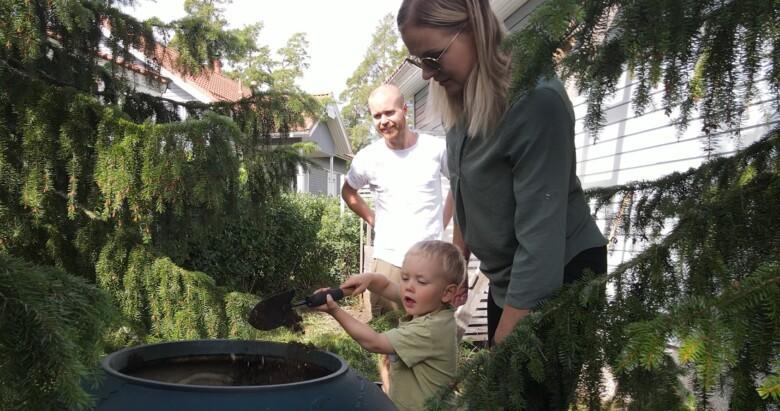 Perhe kompostointi-astian ympärillä. Lapsi laittaa puutarhalapiolla kuiviketta kompostiin.
